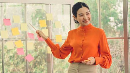 Interior, a teacher gestures to a board and post-it notes, for “How to Talk to your Clients about Investing in Alternative Assets